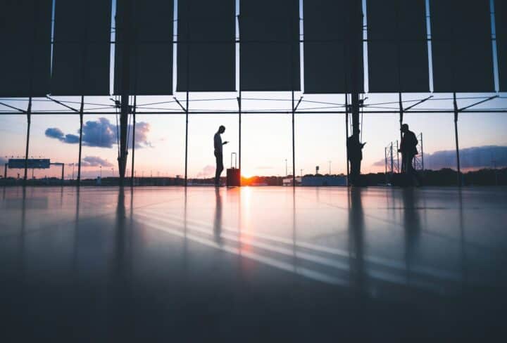 Image of an airport departure area with travellers silhouettes against a setting sun that can be seen through the window