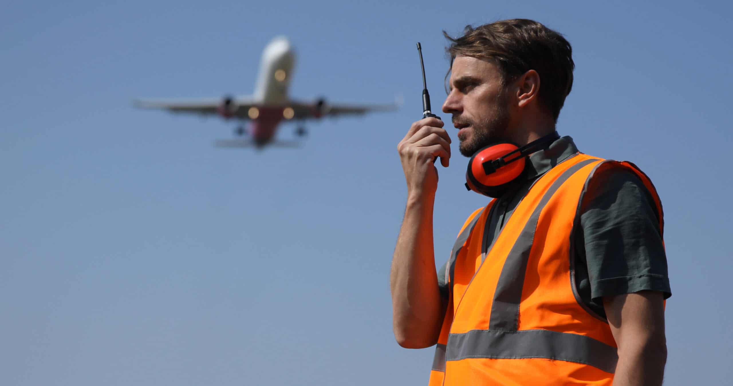 Image of a ground staff worker on a runway talking into a radio while an airliner flies overhead