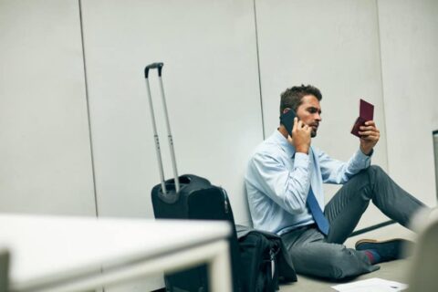 Image of a young man sitting on the floor of an airport. He is looking at his passport while on the phon