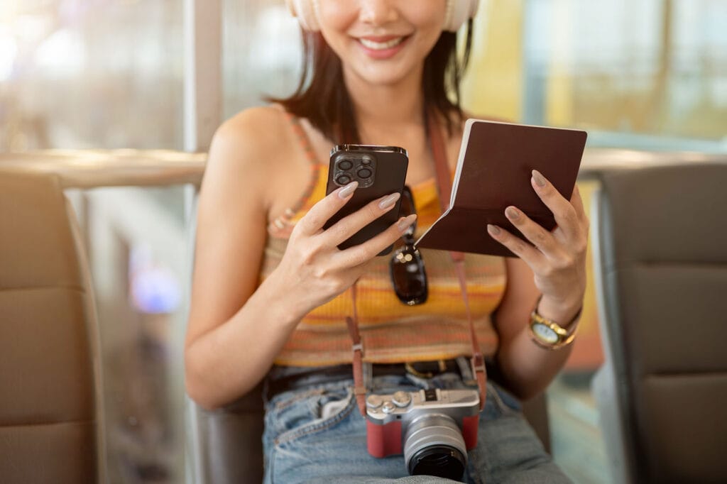 Een jonge vrouw in de vertrekhal van een luchthaven controleert haar vluchtgegevens op een mobiele app terwijl ze haar paspoort vasthoudt